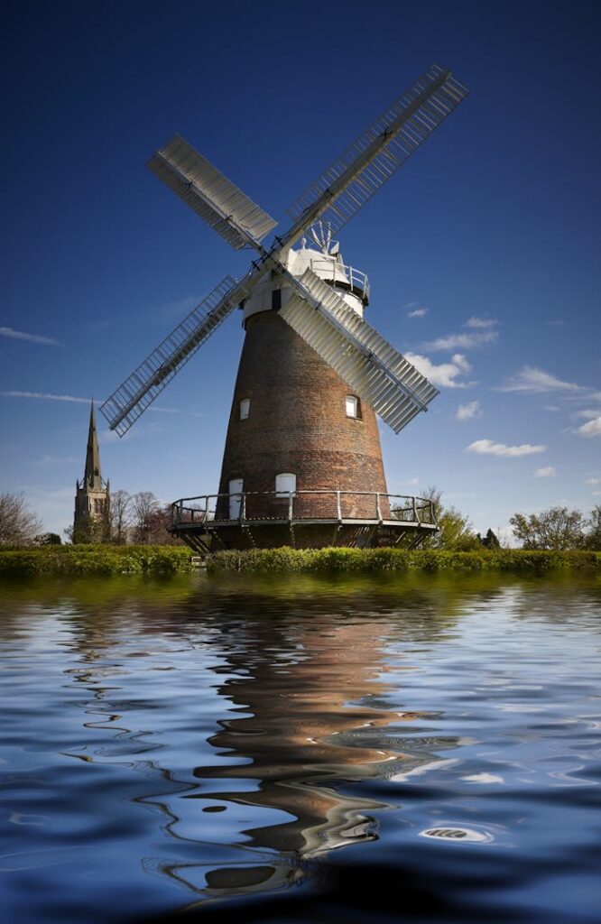 Charming windmill and church reflected in a serene riverside setting under a clear blue sky.