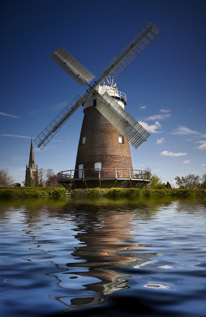 Charming windmill and church reflected in a serene riverside setting under a clear blue sky.